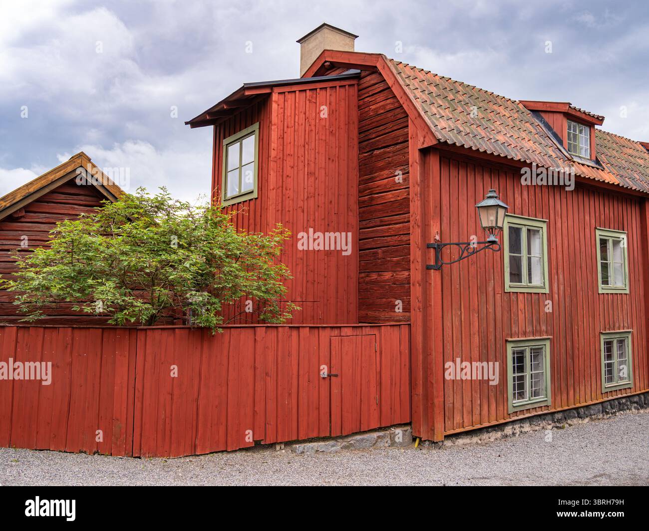 Sodertalje, Svezia - 6 giugno 2025: Un'affascinante casa in legno rosso con tetto inclinato e finestre verdi, circondata da un sentiero di ghiaia e un piccolo autobus Foto Stock
