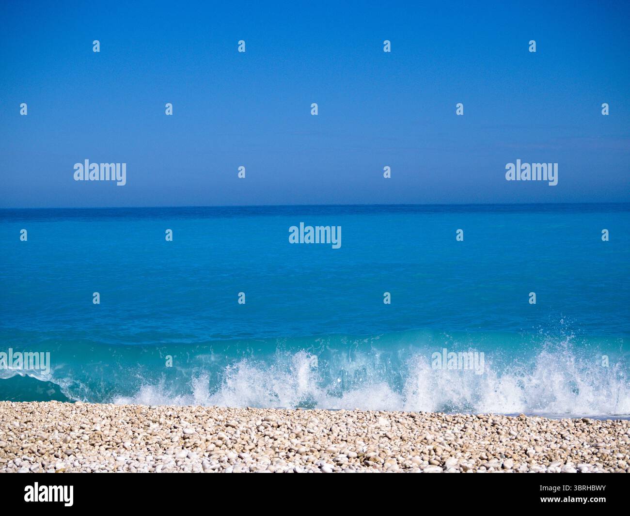 Spiaggia minimalista con ghiaia bianca, onde morbide e orizzonte dove il cielo blu si fonde con il mare. Foto Stock
