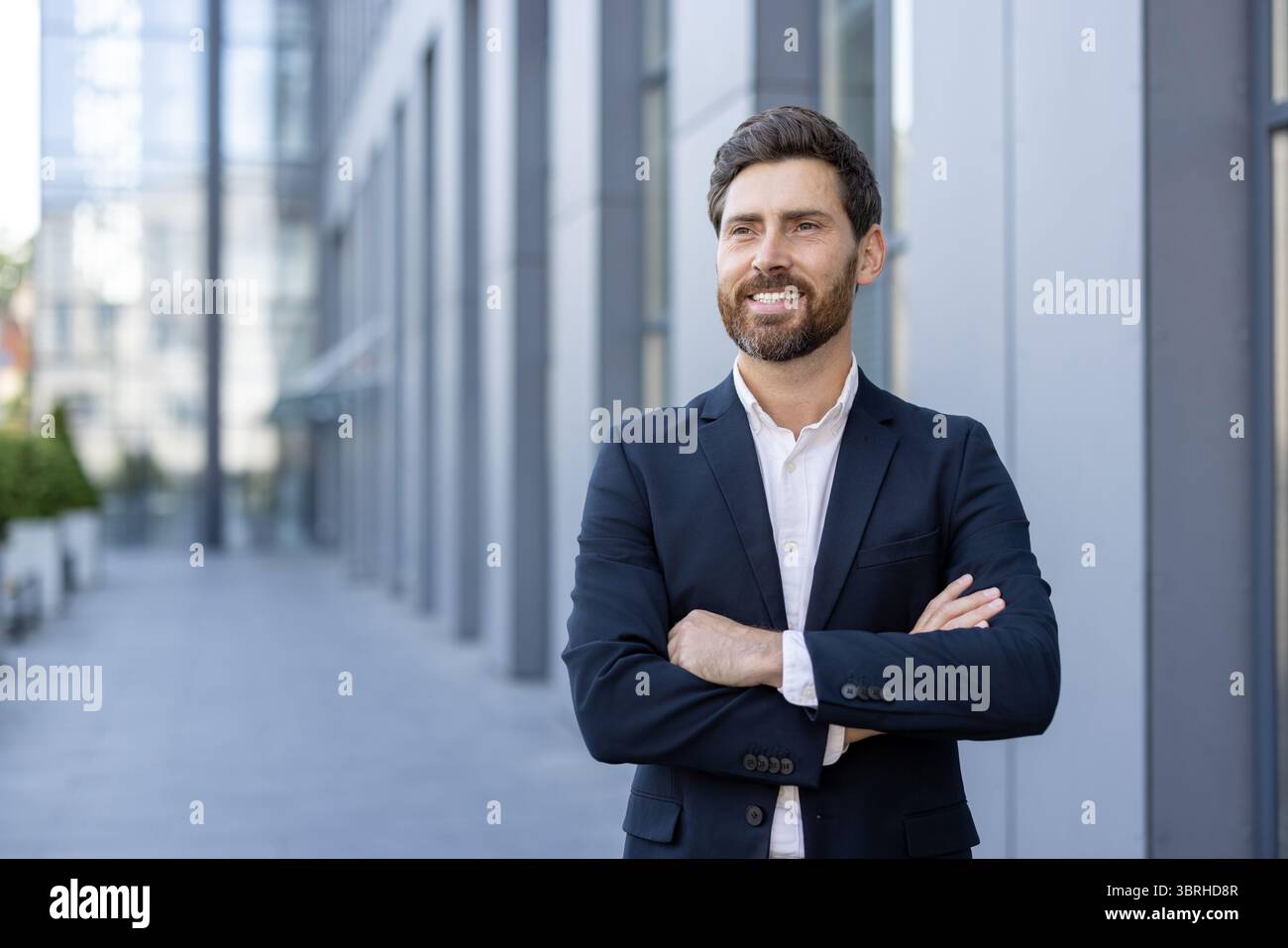 Un uomo d'affari sorridente si trova con sicurezza all'esterno, con le braccia incrociate, di fronte a un edificio moderno, trasuda successo professionale e acume d'affari. Foto Stock