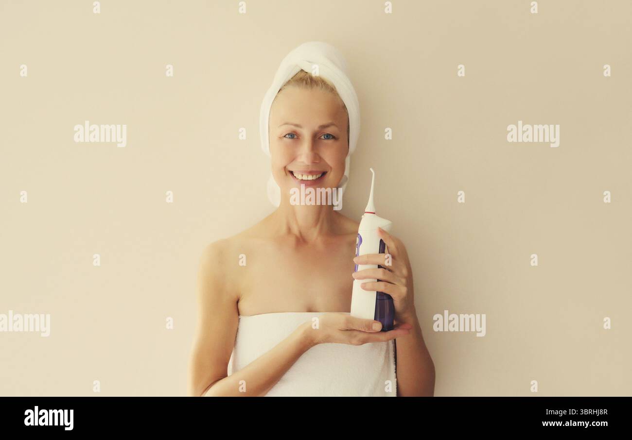 Ritratto di una donna caucasica di mezza età sorridente e felice con denti bianchi e puliti tiene l'irrigatore orale e si asciuga i capelli bagnati con un asciugamano avvolto sul cigno Foto Stock