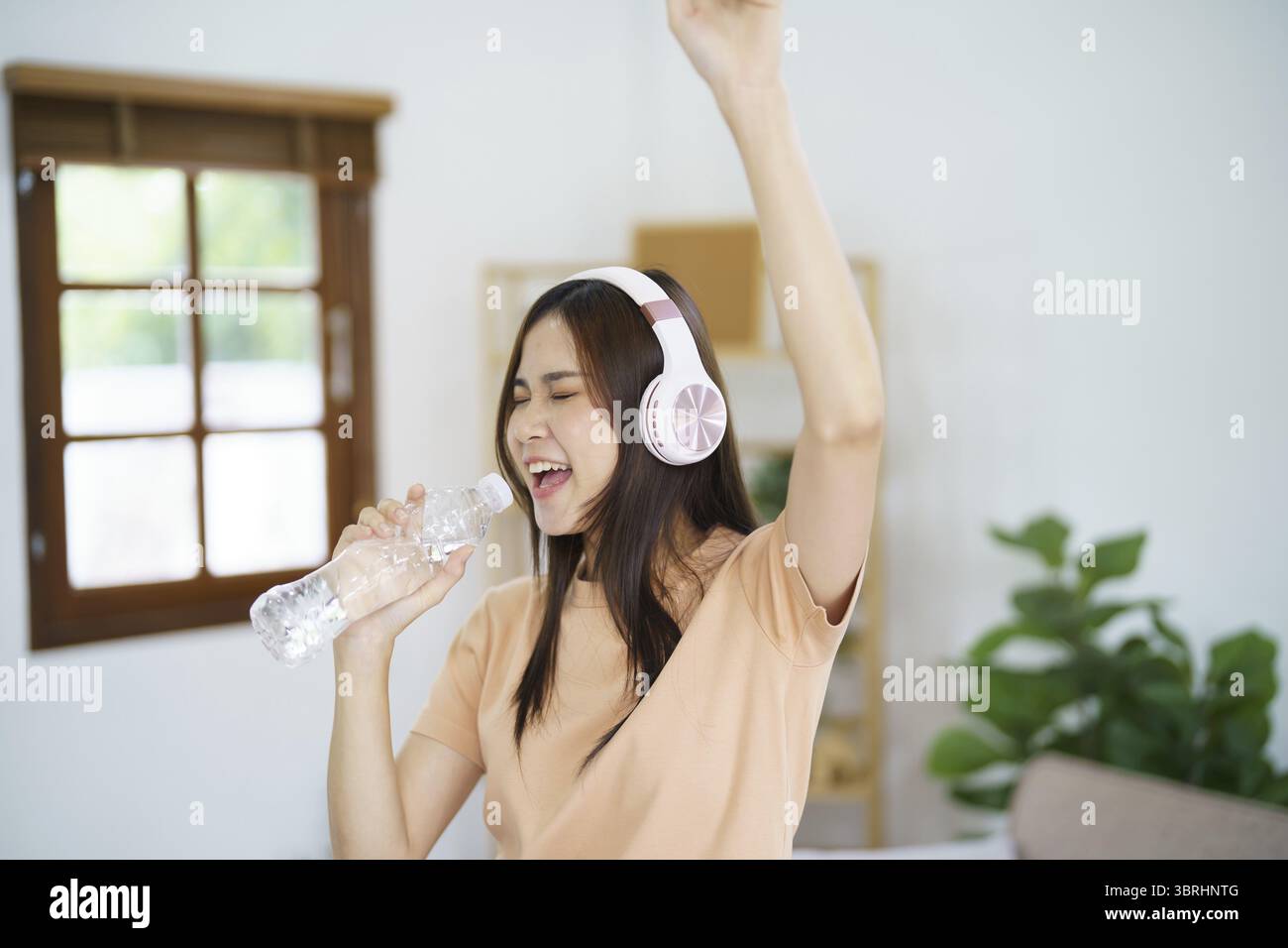 Felice giovane donna asiatica che canta da sola con il microfono a bottiglia e indossa le cuffie ama la musica buon relax in salotto Foto Stock