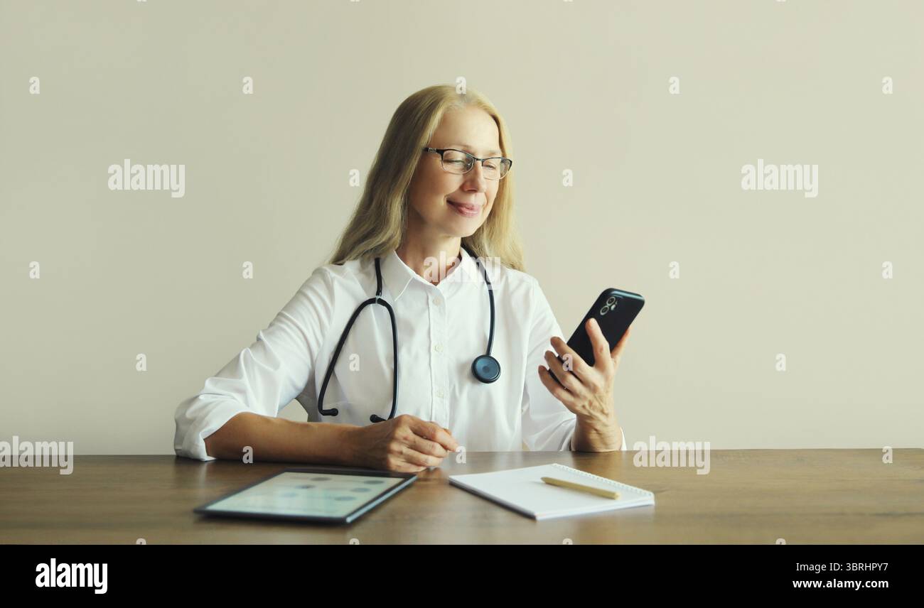 Medico caucasico di mezza età che usa il telefono cellulare con documenti cartacei e tablet digitale che siede alla scrivania nell'ufficio della clinica Foto Stock