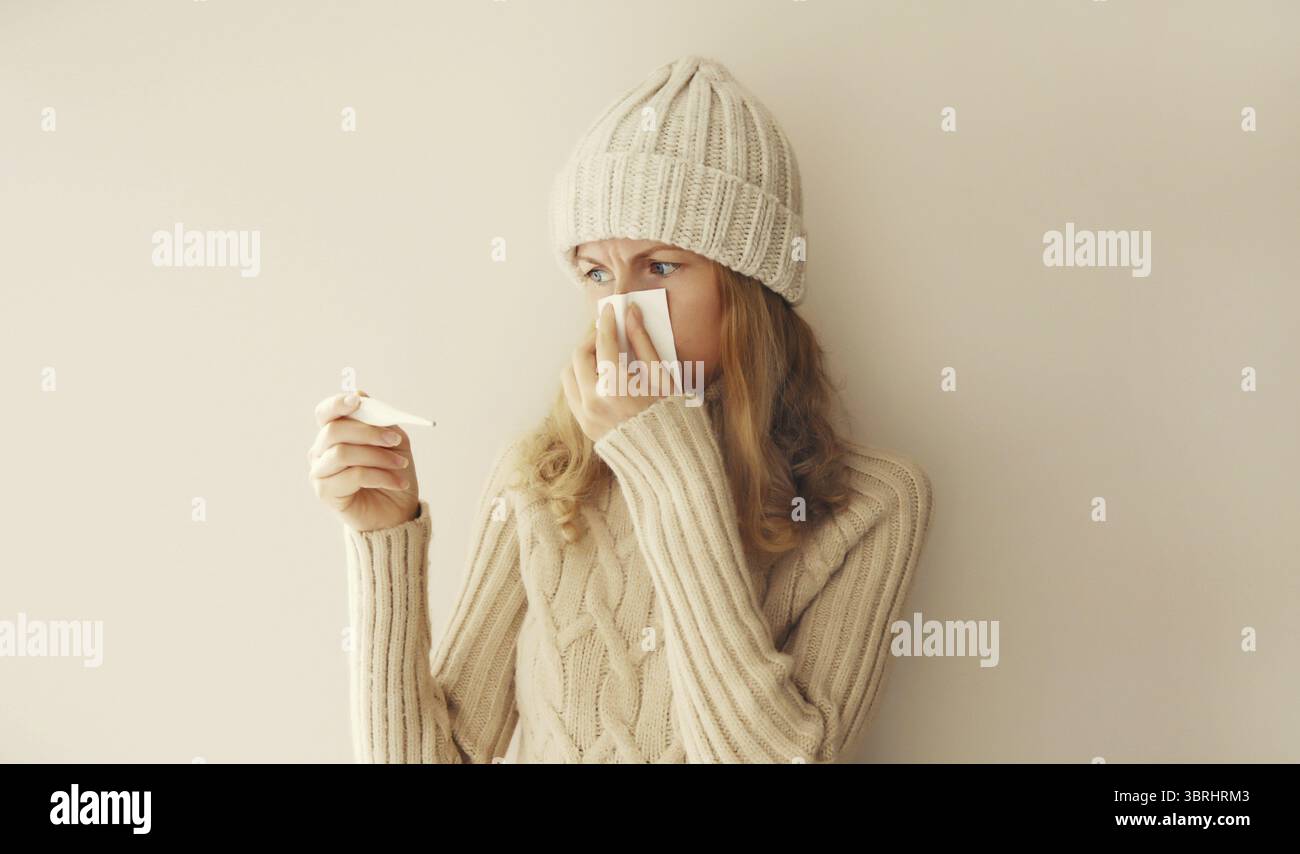 Donna malata che starnutisce il naso soffiato usando il tessuto e misurando la temperatura corporea guardando il termometro indossando abiti caldi e morbidi in maglia, cappello e S Foto Stock