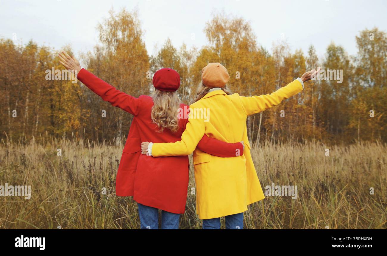 Vista sul retro, due giovani amiche felici nel parco autunnale che si godono il caldo tempo, eleganti fidanzate che alzano le mani insieme indossando il giallo rosso co Foto Stock