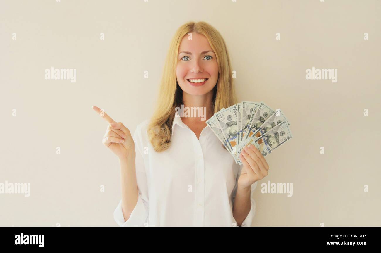 Felice sorridente giovane donna che tiene in mano soldi in contanti in banconote da un dollaro e punta il dito su un lato, spazio vuoto per la copia del testo pubblicitario Foto Stock