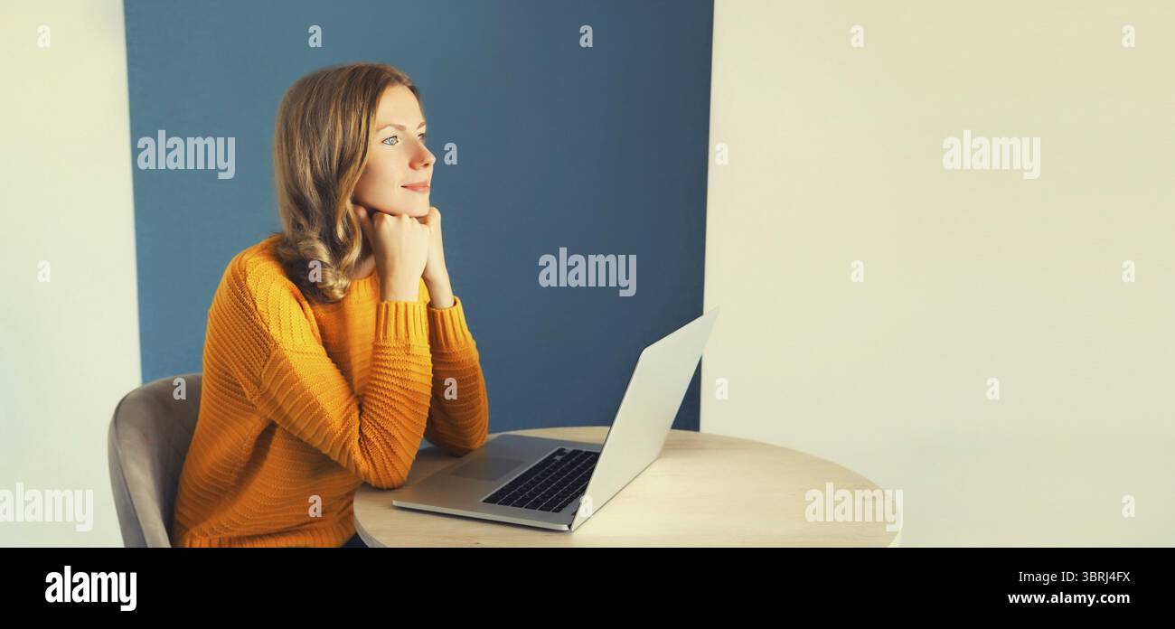 Giovane donna moderna che lavora con un computer portatile seduto alla scrivania in camera a casa e che guarda fuori dalla finestra Foto Stock