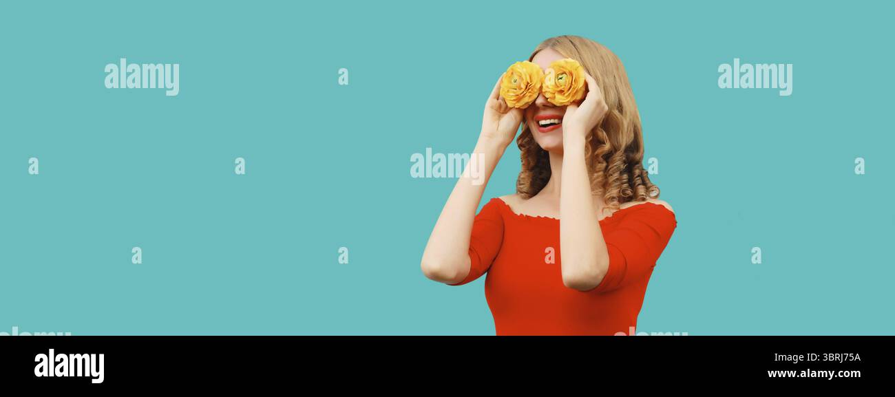 Ritratto estivo di una bella donna sorridente che copre gli occhi di gemme di fiori come binocolo alla ricerca di qualcosa sullo sfondo blu di uno studio Foto Stock