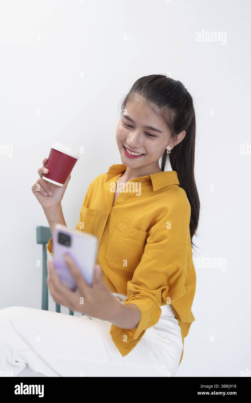 Donna asiatica che tiene il telefono e la tazza di caffè con camicia gialla su sfondo bianco Foto Stock
