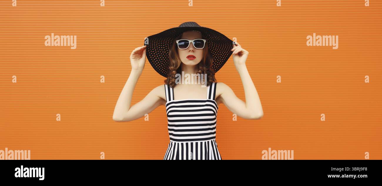 Ritratto di una bella giovane donna che posa indossando un cappello di paglia estivo nero e un vestito su sfondo arancione Foto Stock
