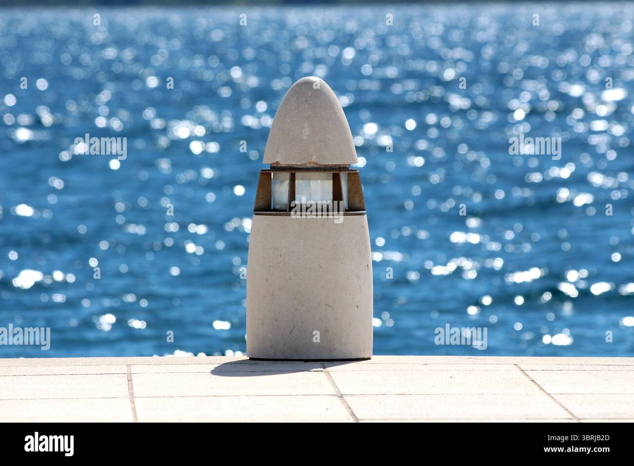 Una robusta lampada in pietra con cappuccio conico e centro in vetro si erge su un pavimento piastrellato, sullo sfondo di acqua blu scintillante Foto Stock