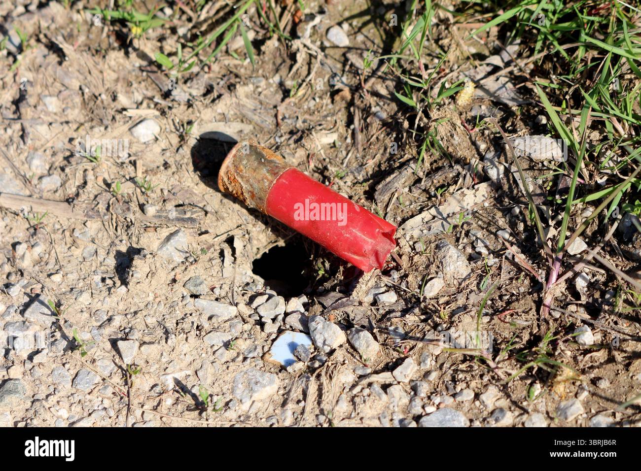 Un involucro rosso intemprato del fucile si trova abbandonato nello sporco e nella ghiaia, parzialmente sepolto e arrugginito, evidenziando l'inquinamento ambientale Foto Stock
