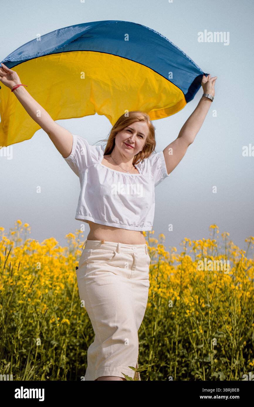 Un gesto simbolico di orgoglio nazionale come donna ondeggia la bandiera dell'Ucraina nei pressi di un campo di colza fiorito il giorno della Costituzione Foto Stock