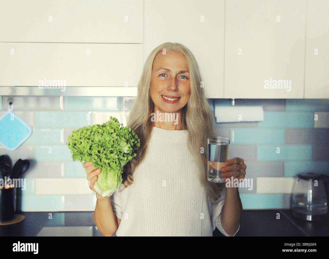 Ritratto di una donna dai capelli grigi di mezza età sorridente sorridente sorridente che tiene in cucina la mattina presto a casa lattuga verde e bicchiere con acqua fresca e pulita Foto Stock Ritratto di una donna dai capelli grigi di mezza età sorridente sorridente sorridente che tiene in cucina la mattina presto a casa lattuga verde e bicchiere con acqua fresca e pulita Foto Stock