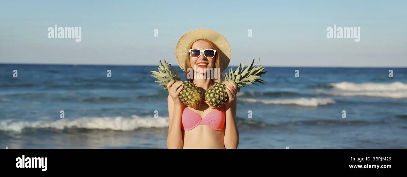 Vacanze estive, bella donna sorridente e felice con costume da bagno bikini rosa, cappello di paglia turistico che regge frutti di ananas sulla spiaggia sulla costa del mare Foto Stock