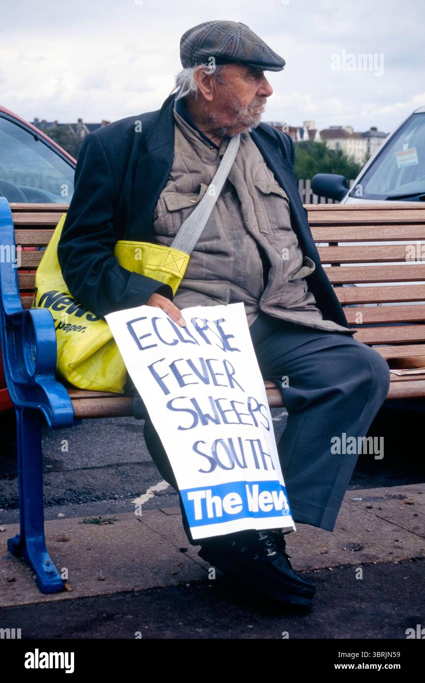 il venditore di giornali anziani sedeva sul panchina sul lungomare tenendo un grande poster scritto a mano con titoli di coda la febbre dell'eclissi spazza verso sud riferendosi all'eclissi solare totale agosto 1999 inghilterra regno unito Foto Stock