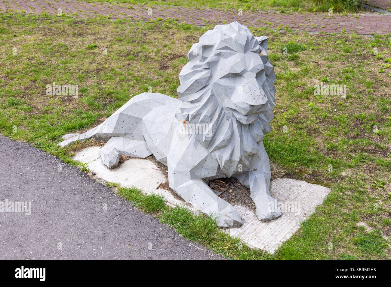 Una scultura di leone geometrica e angolare in cemento poggia su una lastra di cemento tra erba verde e un sentiero lastricato, che mostra arte pubblica moderna. Foto Stock