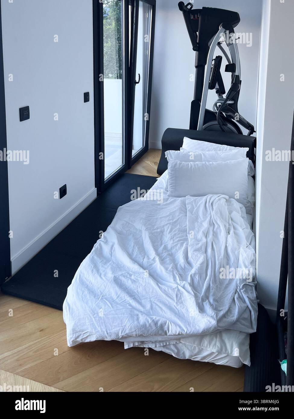 Il letto con biancheria bianca è posizionato in un angolo accogliente, accanto a una moderna macchina per esercizi, che unisce comfort e fitness in un ambiente elegante Foto Stock