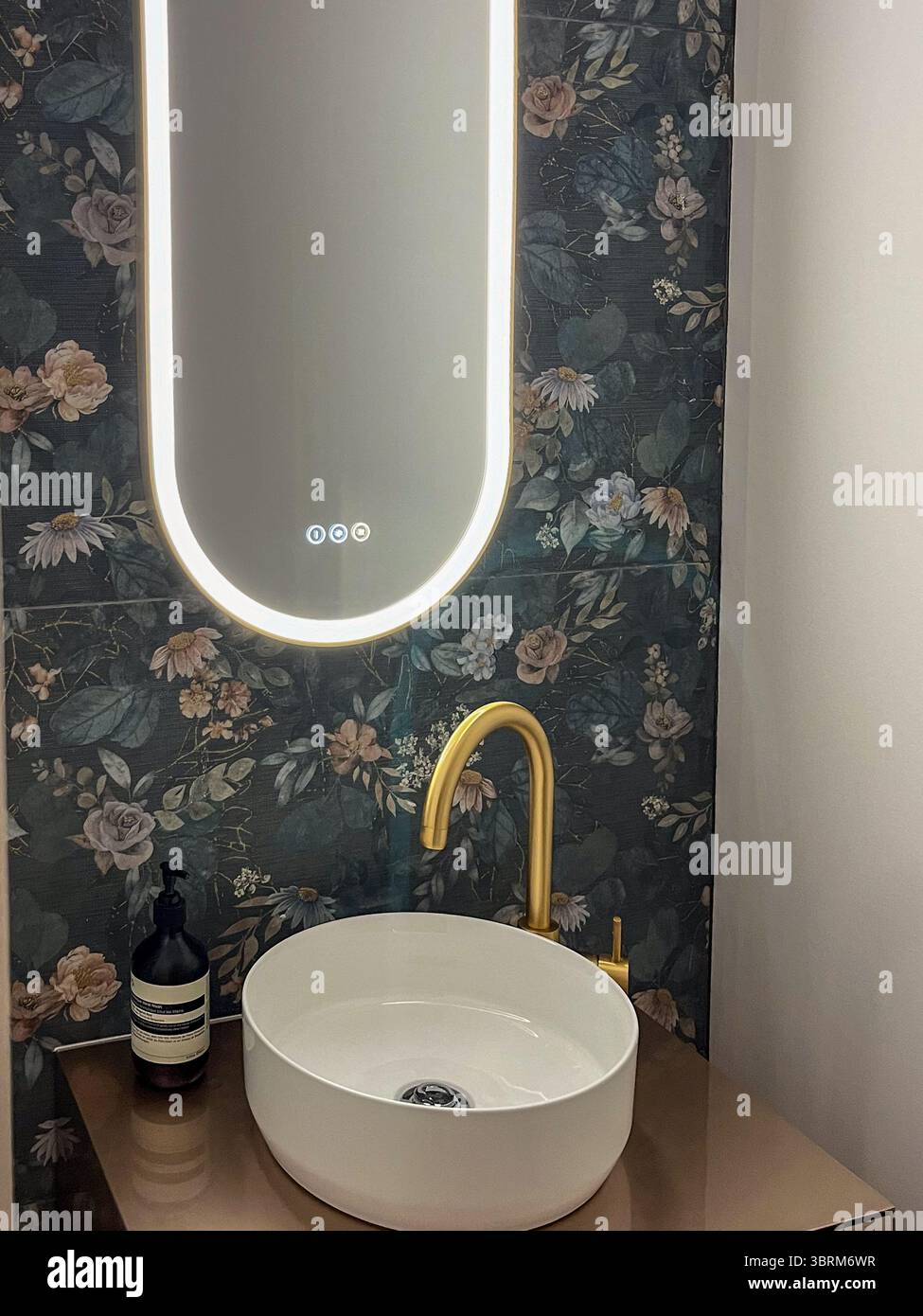 Il bagno dal design contemporaneo presenta un lavandino rotondo con rubinetto dorato, specchio illuminato e carta da parati floreale, che accentuano l'atmosfera tranquilla del Foto Stock