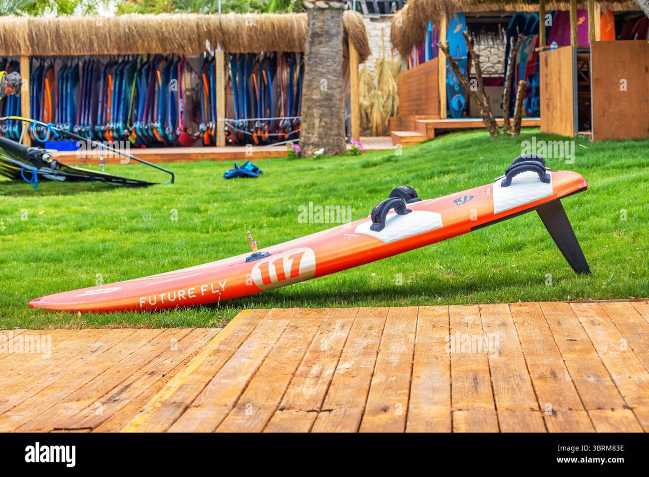 29 aprile 2025, Alacati, Turkiye: Tavola da windsurf arancione sdraiata sull'erba davanti ad una scuola di surf, pronta per la prossima sessione Foto Stock
