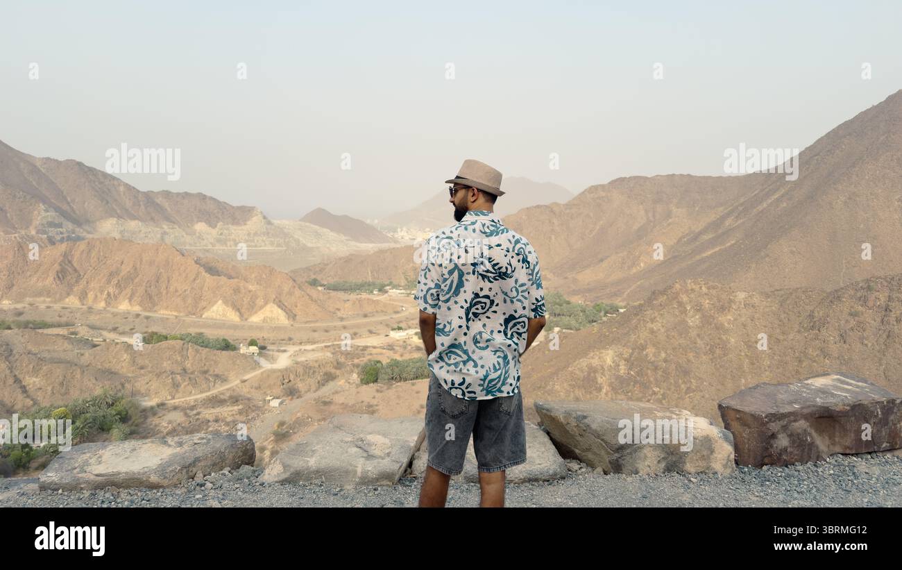Un uomo in piedi su un punto panoramico roccioso, con indosso una camicia a motivi geometrici, pantaloncini e un cappello, che si affaccia su un paesaggio montano panoramico Foto Stock