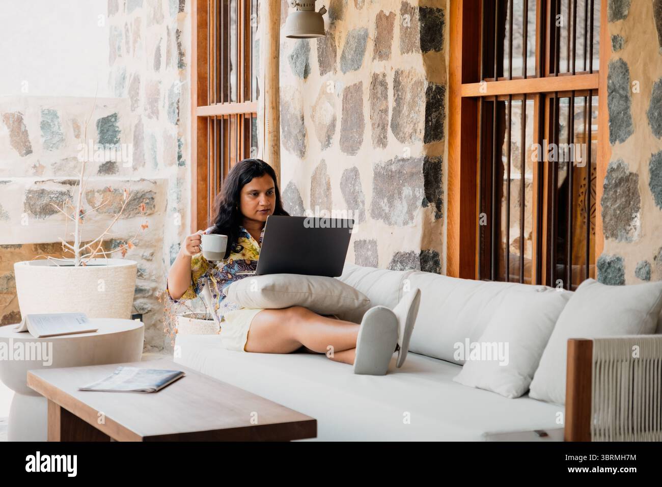 Una donna che si rilassa su un divano del patio, indossa un abito floreale e tiene in mano una tazza, mentre lavora su un notebook di fronte a un muro rustico in pietra Foto Stock