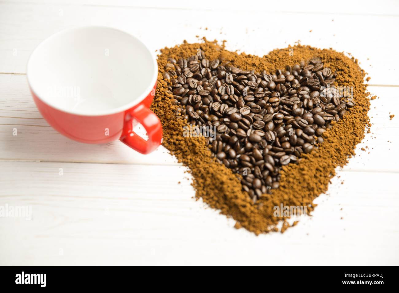 I fondi e i chicchi di caffè a forma di cuore si formano su una superficie piana bianca con una tazza rossa Foto Stock
