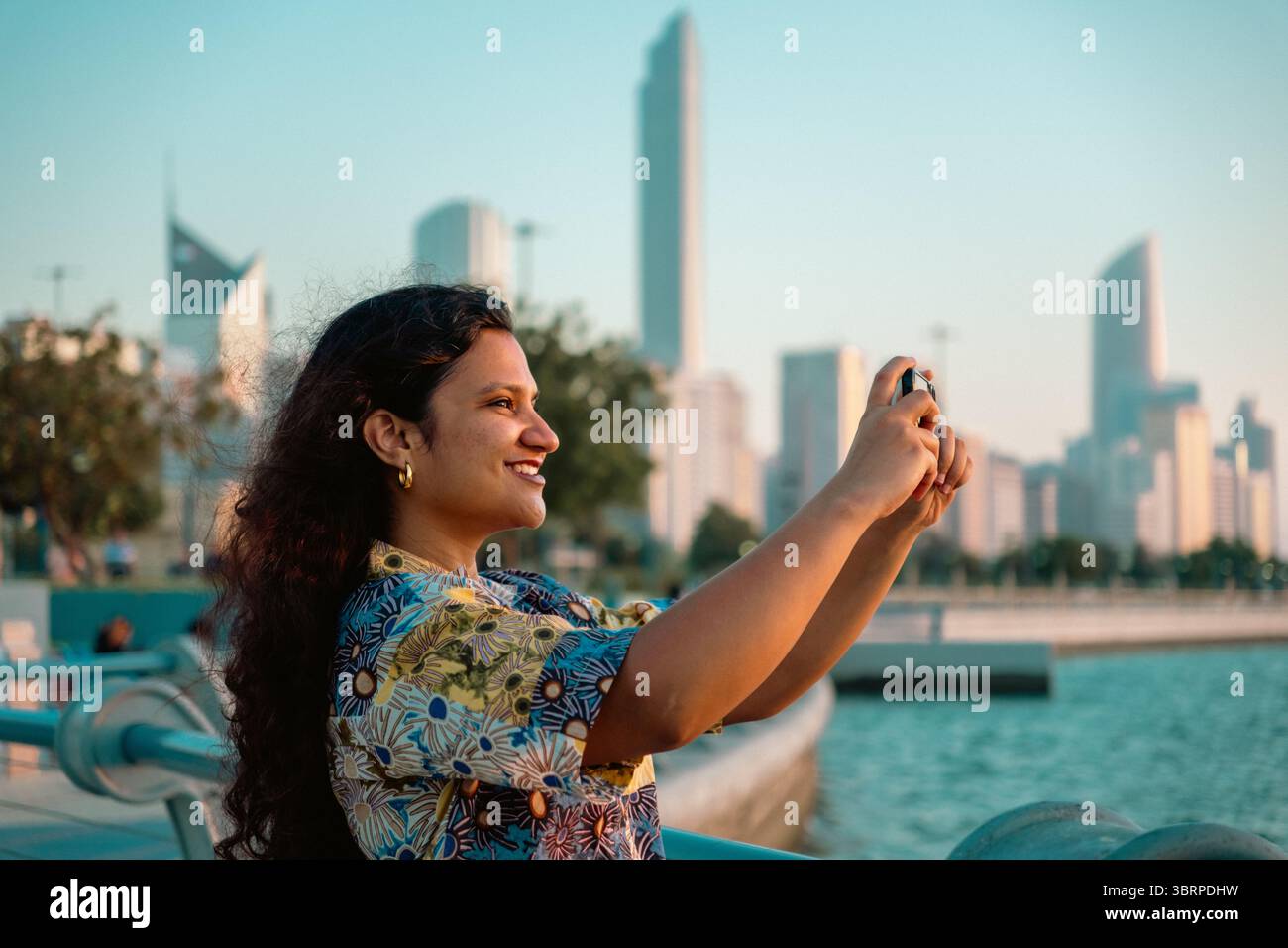 Donna asiatica sorridente che scatta foto della Corniche di Abu Dhabi nel suo smartphone con sfondo skyline cittadino Foto Stock