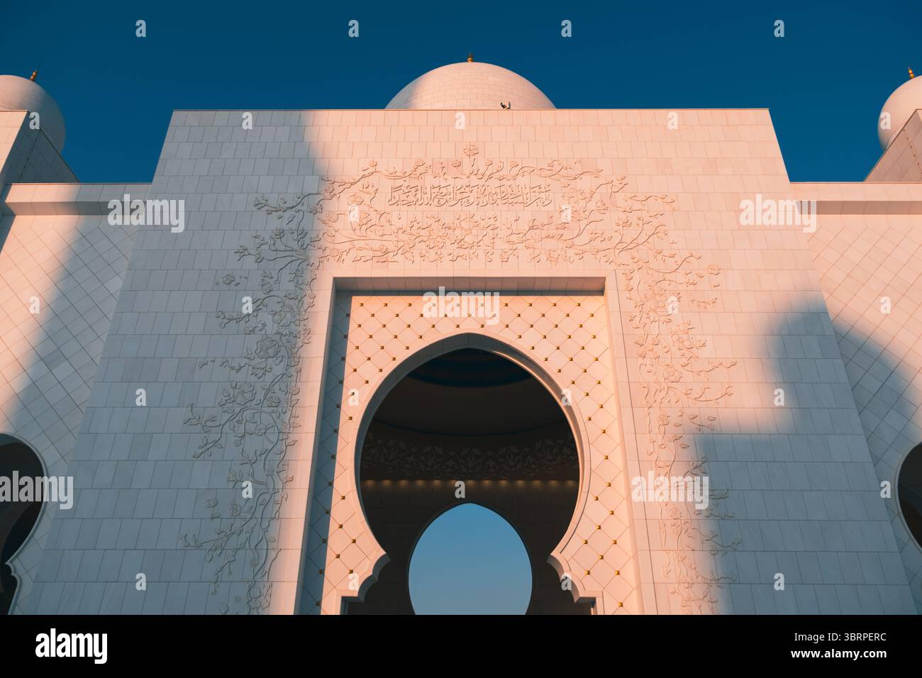 Primo piano dell'arco all'ingresso della grande Moschea dello Sceicco Zayed con intricati motivi floreali e facciata in marmo bianco sotto la luce del tramonto Foto Stock