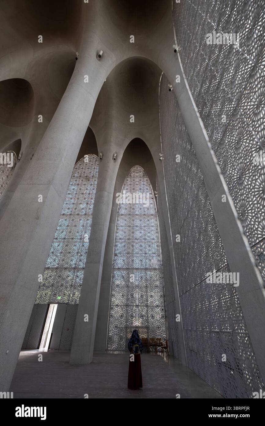 Una donna si erge in una moschea moderna, spaziosa e dal design complesso, con alte colonne ad arco e pareti a motivi geometrici, con un lungo abito e un velo Foto Stock