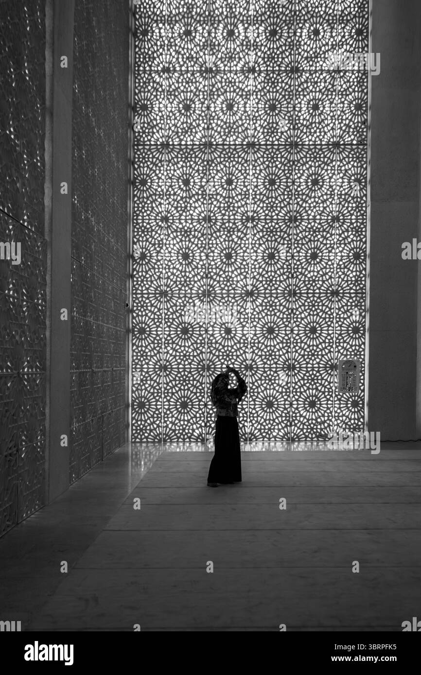 Una persona con un abbigliamento tradizionale si erge di fronte a una parete di vetro con fantasie intricate in un interno spazioso e aperto. Foto Stock