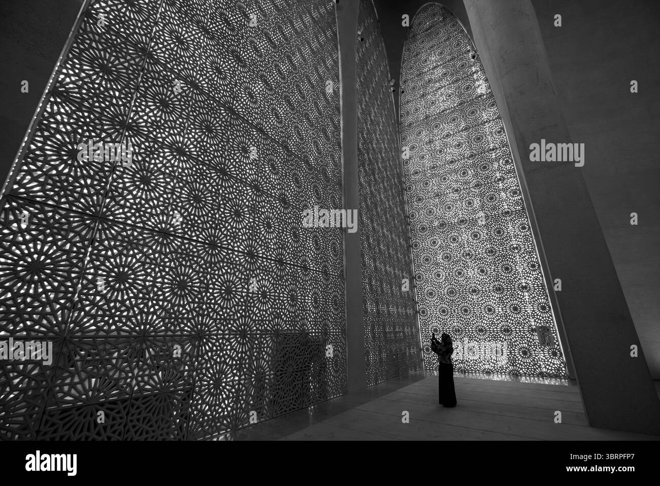 Una donna con un abbigliamento tradizionale che prega all'interno di una grande moschea moderna con pareti intricate e alti soffitti ad arco. Foto Stock