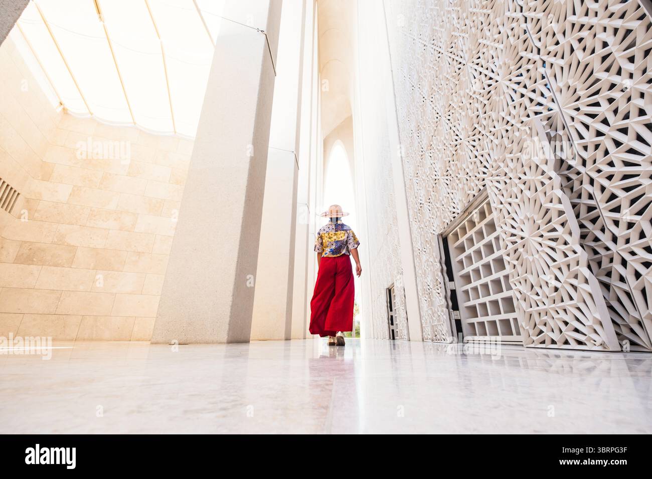 Una donna in abito etnico cammina attraverso uno spazio architettonico con intricati disegni bianchi e alte colonne. Indossa un cappello a tesa larga. Foto Stock