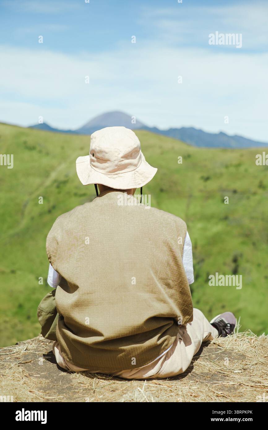 Vista posteriore di un escursionista maschile del sud-est asiatico seduto tranquillamente su una collina erbosa, con indosso un cappello e un giubbotto leggero, godendosi le verdi colline Foto Stock