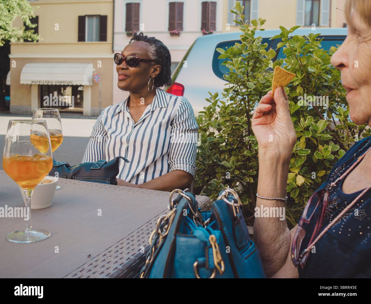 Due donne mature che si godono un momento di relax insieme, bevono aperitivi e mangiano patatine in un caffè all'aperto, conversano e si divertono Foto Stock