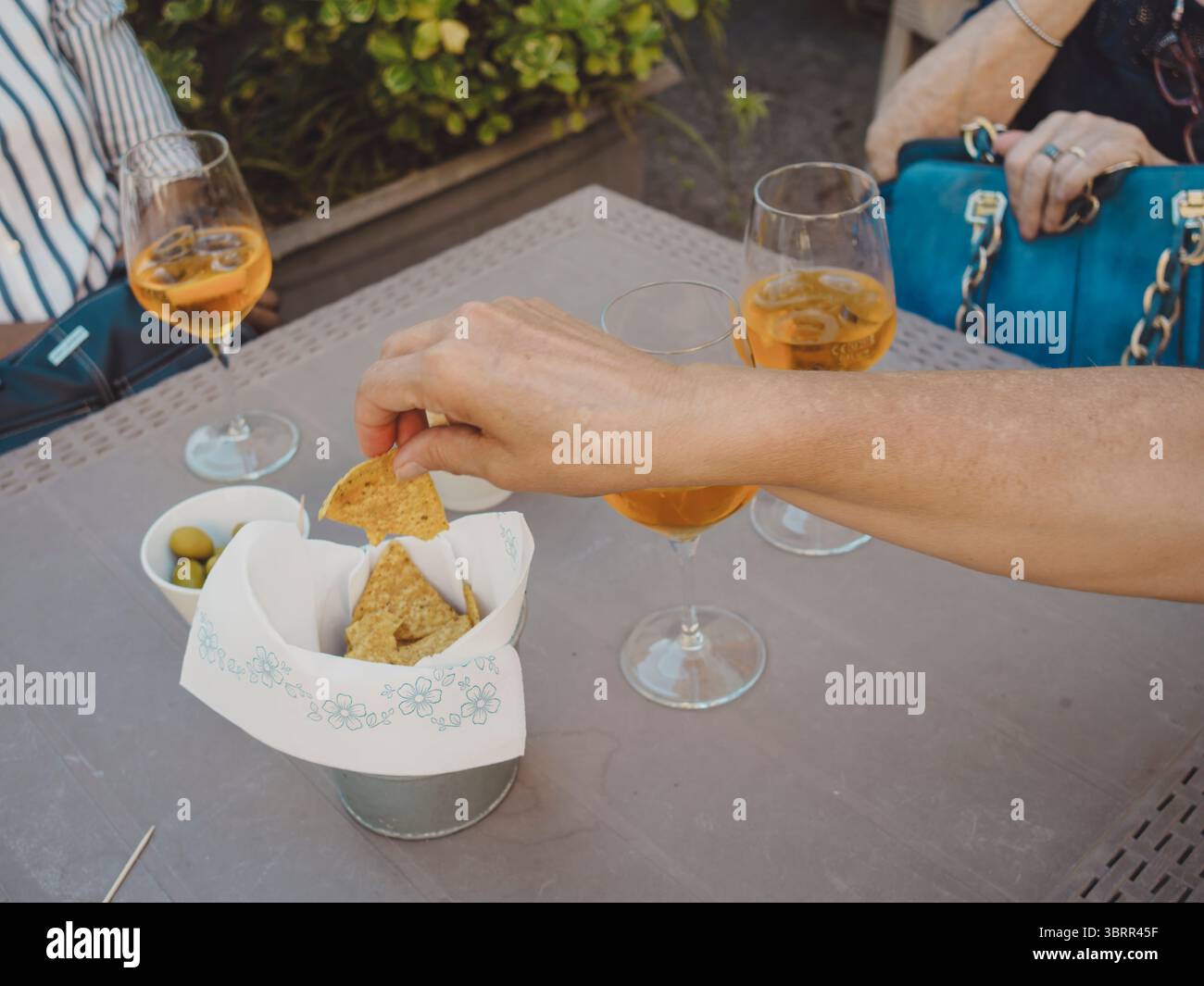 Donna caucasica adulta che raccoglie nachos da un secchio di metallo mentre beve un aperitivo all'arancia con gli amici a un tavolo del bar, con olive verdi e altri gl Foto Stock