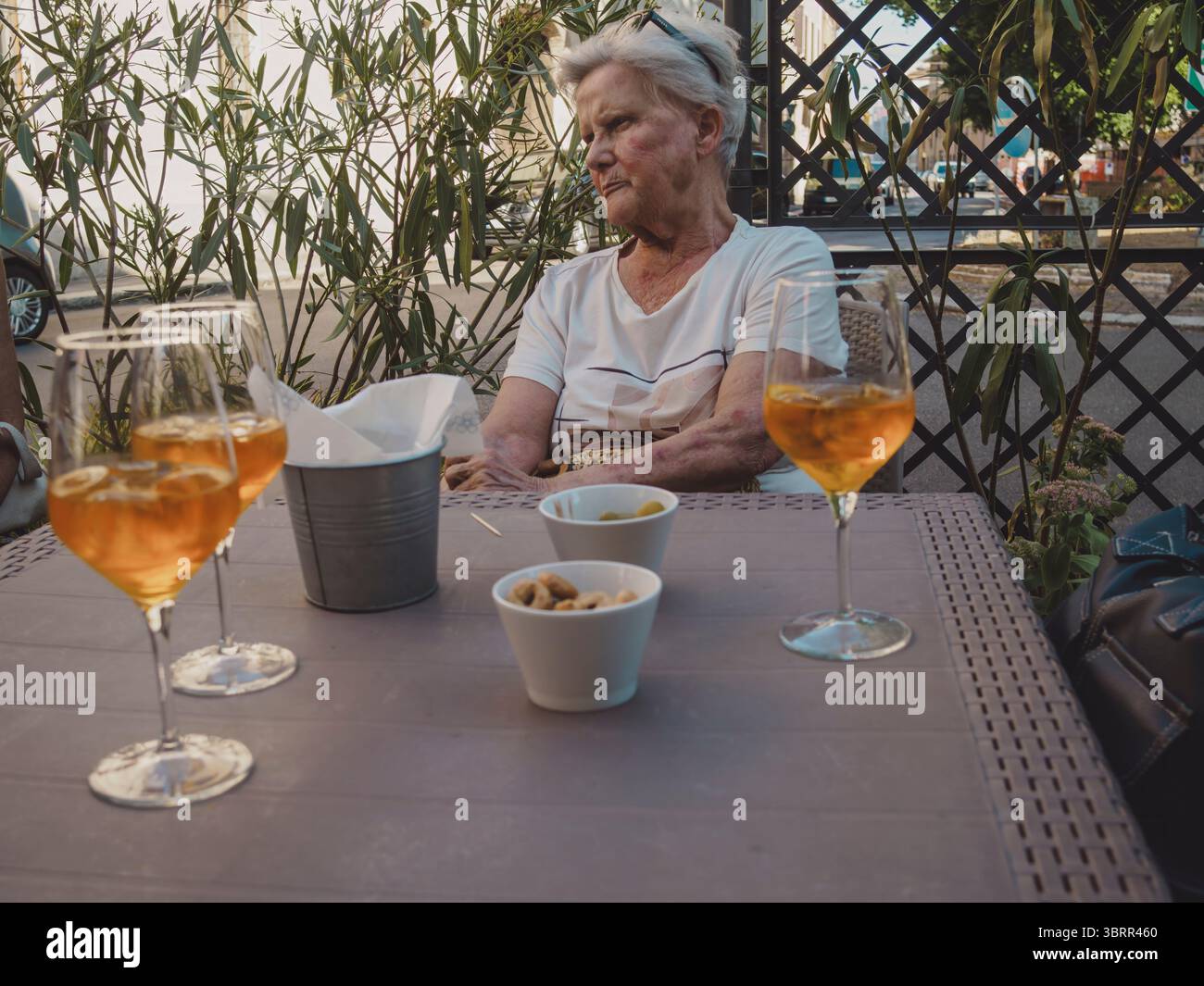 Anziana donna caucasica con capelli bianchi seduta ad un tavolino all'aperto, gustando un rinfrescante aperitivo con spuntini, circondato da piante Foto Stock