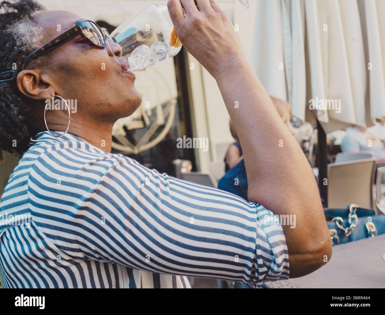 Elegante donna afroamericana senior con occhiali da sole e grandi orecchini a cerchio, con camicia a righe, si gode un cocktail rinfrescante in un bar all'aperto Foto Stock
