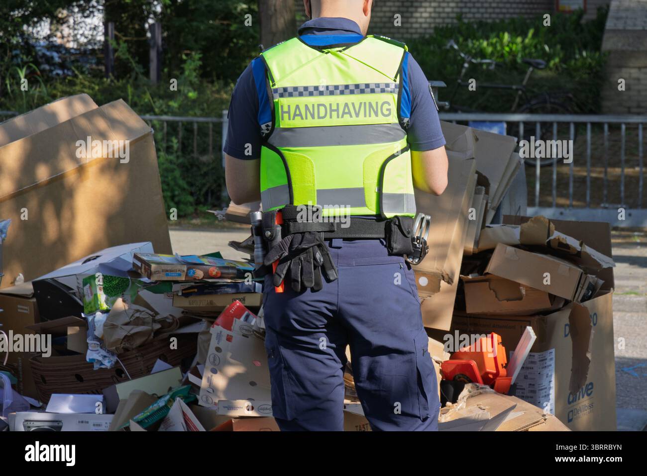 Un ufficiale di "Handhaving" (applicazione) in un giubbotto riflettente si trova in mezzo a una pila di cartone e spazzatura scartati, suggerendo la gestione dei rifiuti o il pubblico Foto Stock