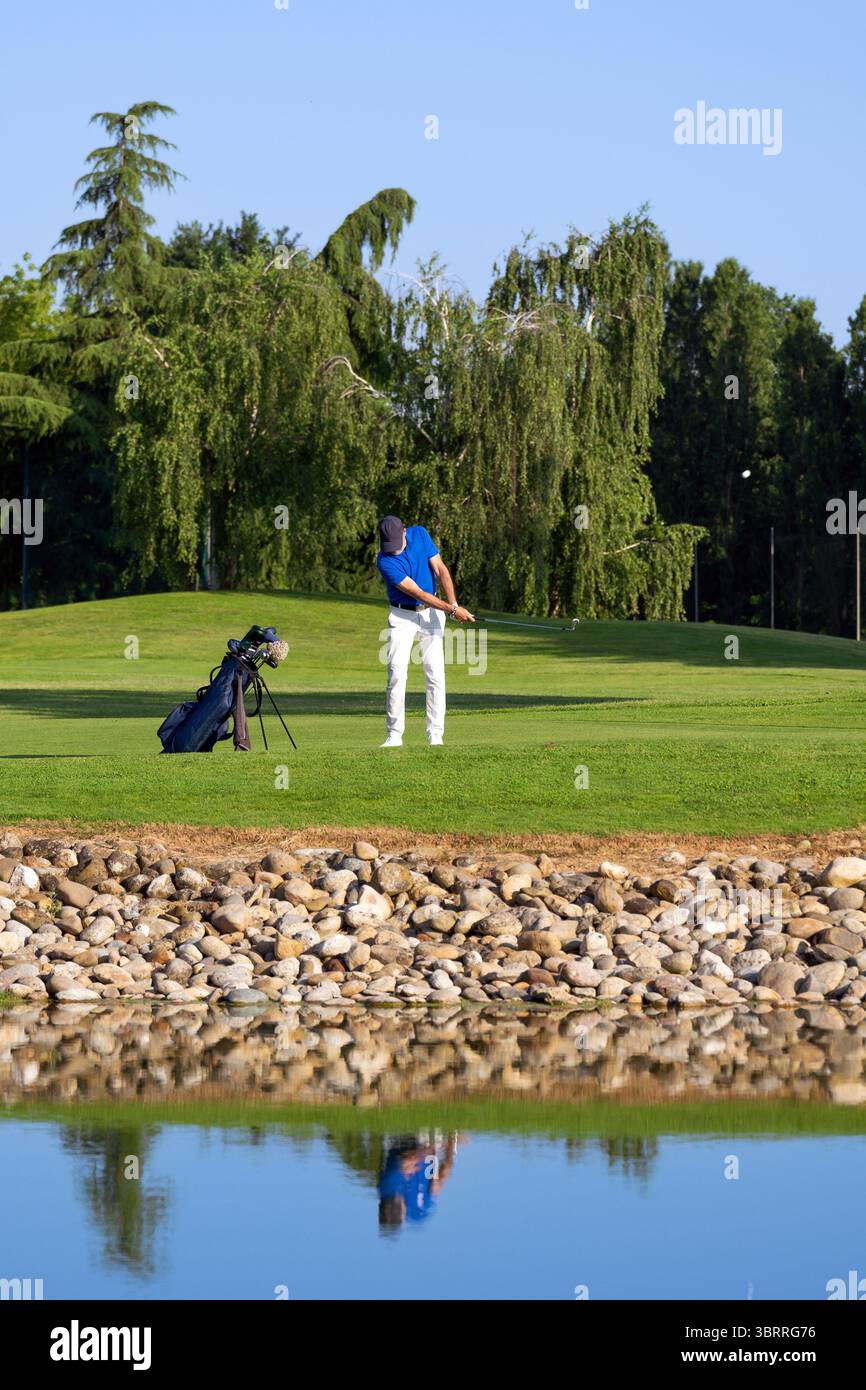 Golfista che batte la palla in una bella giornata di sole con alberi e laghetto sullo sfondo Foto Stock