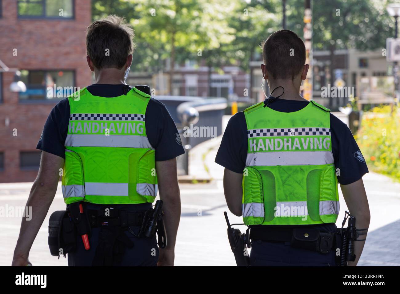 Due ufficiali di polizia, visti da dietro, indossano giubbotti riflettenti, camminano per una strada di Utrecht, Paesi Bassi. Foto Stock