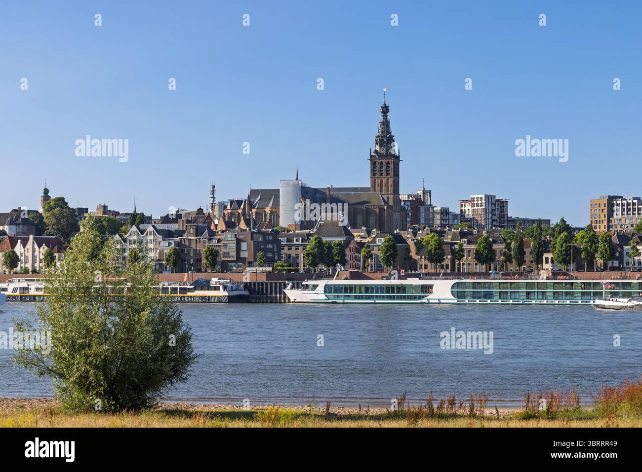 La magnifica guglia della chiesa di Stevenskerk domina lo storico skyline di Nijmegen, Paesi Bassi, mentre le navi da crociera sul fiume attraccano lungo il Waal, tutte bagnate Foto Stock