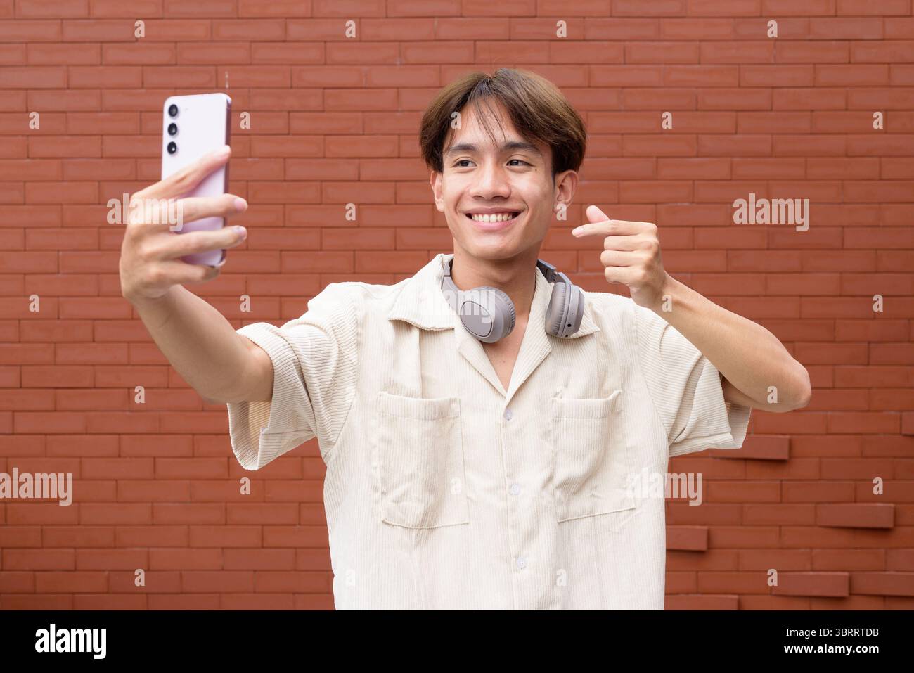 Ritratto di non binario carino asiatico giovane gay uomo contro muro di mattoni usando il telefono Foto Stock
