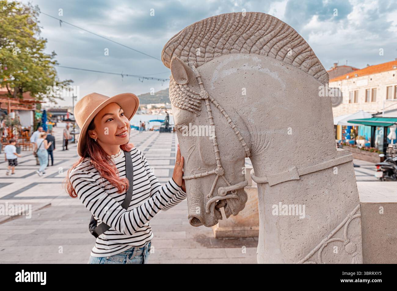7 maggio 2025, Foca, Turkiye: Giovane donna con cappello e camicia a righe che ammira una scultura di cavallo di pietra in una piazza storica Foto Stock