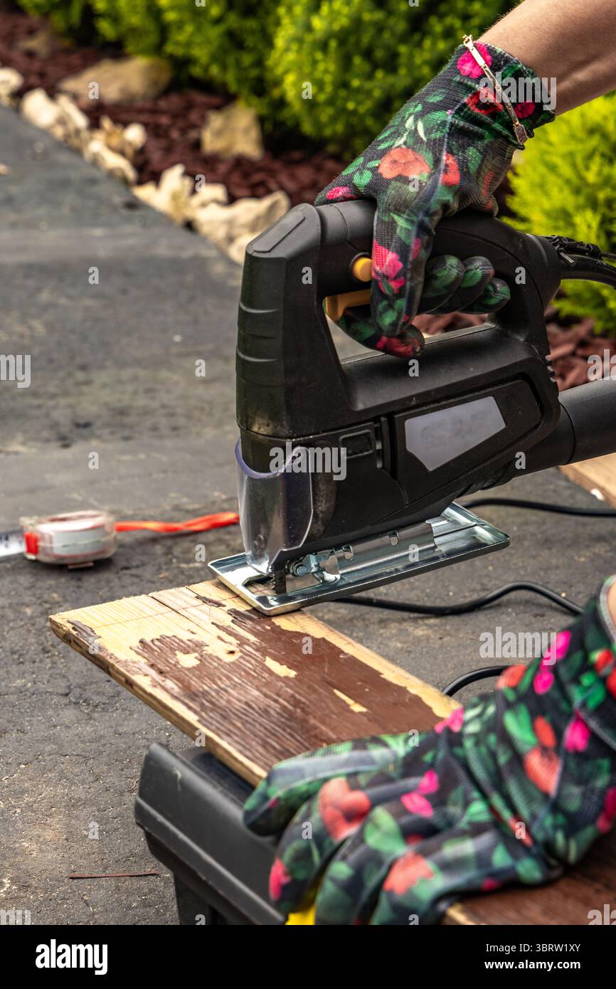 Una donna che indossa guanti protettivi colorati taglia le tavole in giardino utilizzando un seghetto elettrico. Taglieri per un ponte giardino, costruzione di una veranda, o Foto Stock