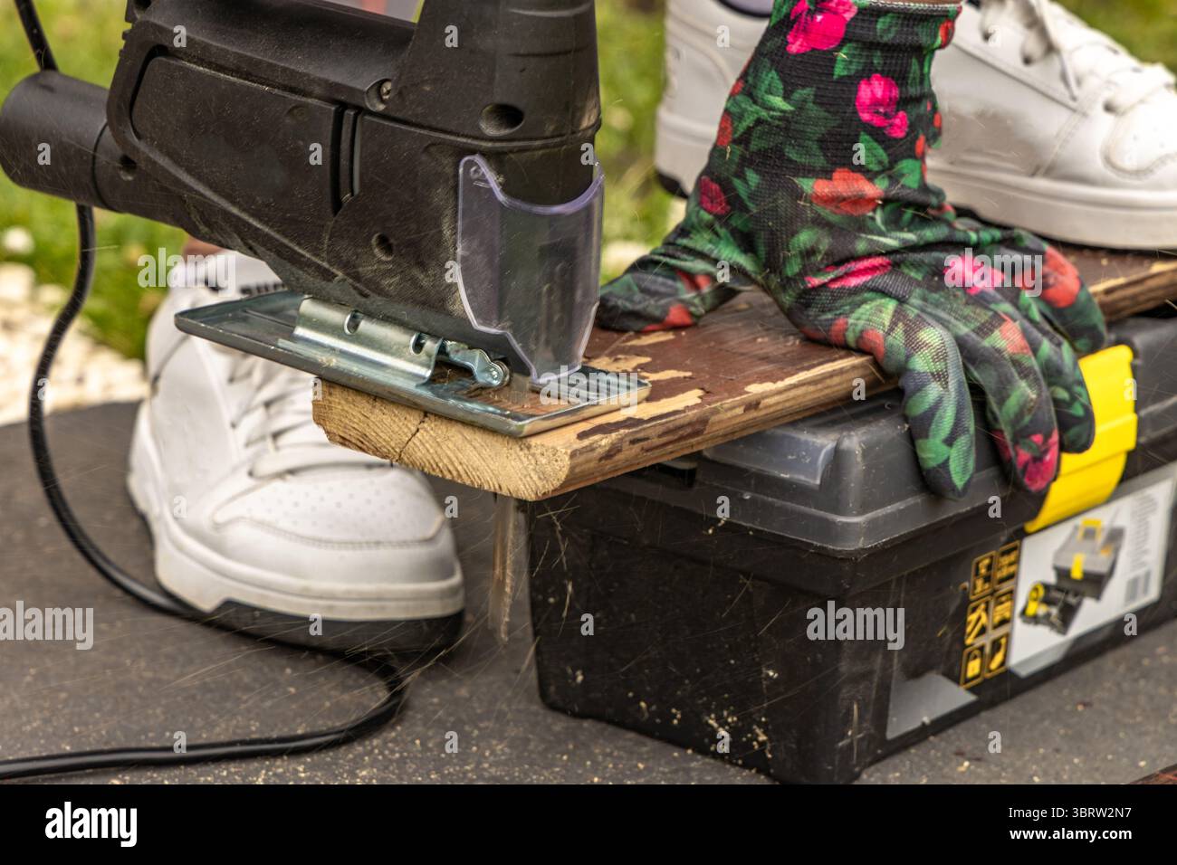 Una donna che indossa guanti protettivi colorati taglia le tavole in giardino utilizzando un seghetto elettrico. Taglieri per un ponte giardino, costruzione di una veranda, o Foto Stock
