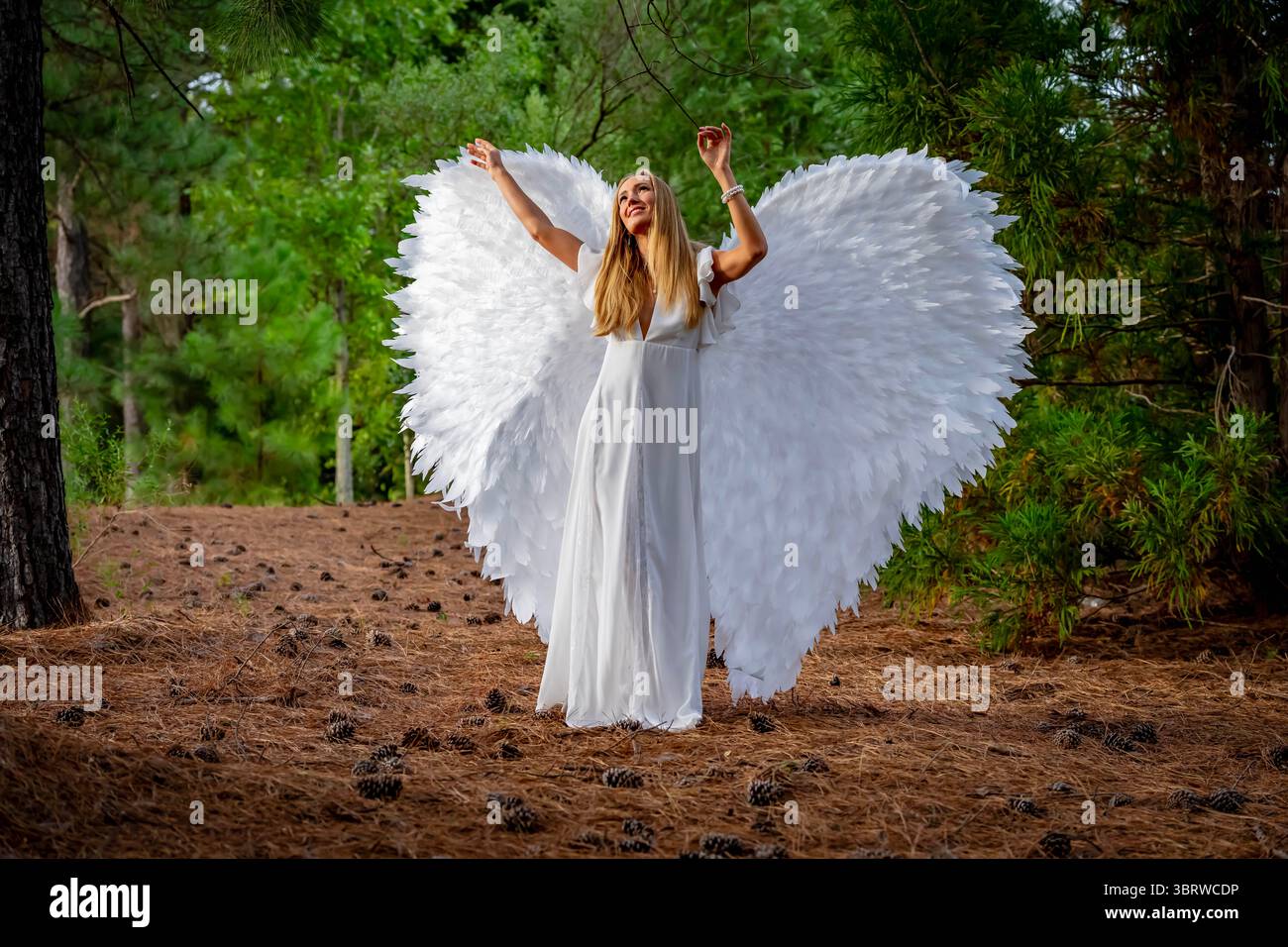 Una splendida giovane bionda vestita di bianco puro, adornata da delicate ali d'angelo, crea una posa graziosa. La sua espressione serena e il suo abbigliamento fluido ev Foto Stock