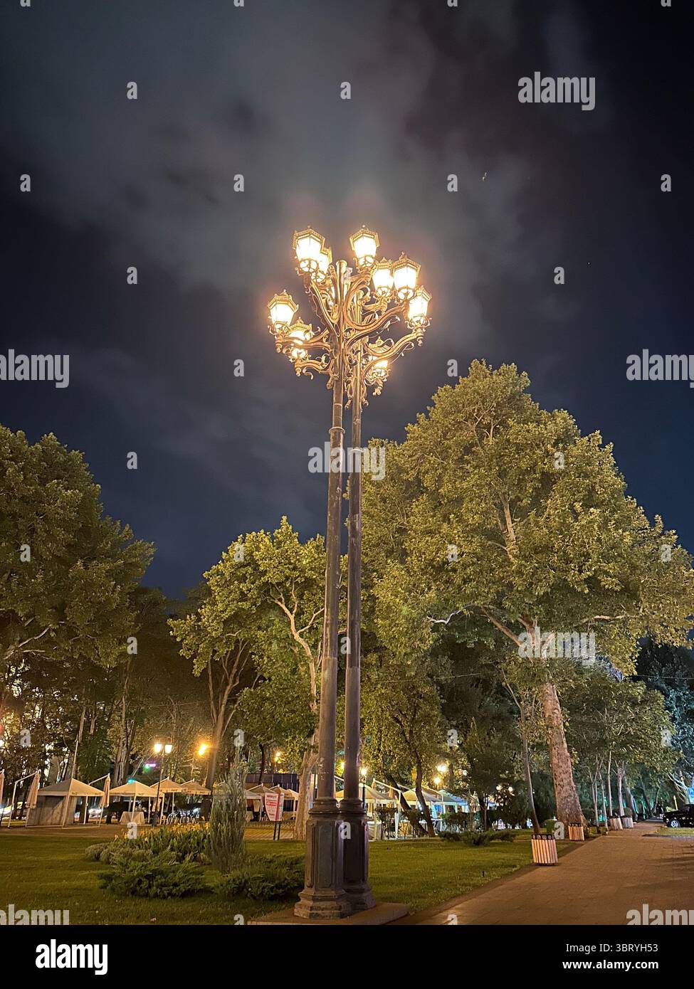 Un lampione a più teste splendidamente ornato che si illumina calorosamente in un tranquillo parco urbano di notte. Circondato da alti alberi e tende da un mercato. - Immagine stock catturata con smartphone