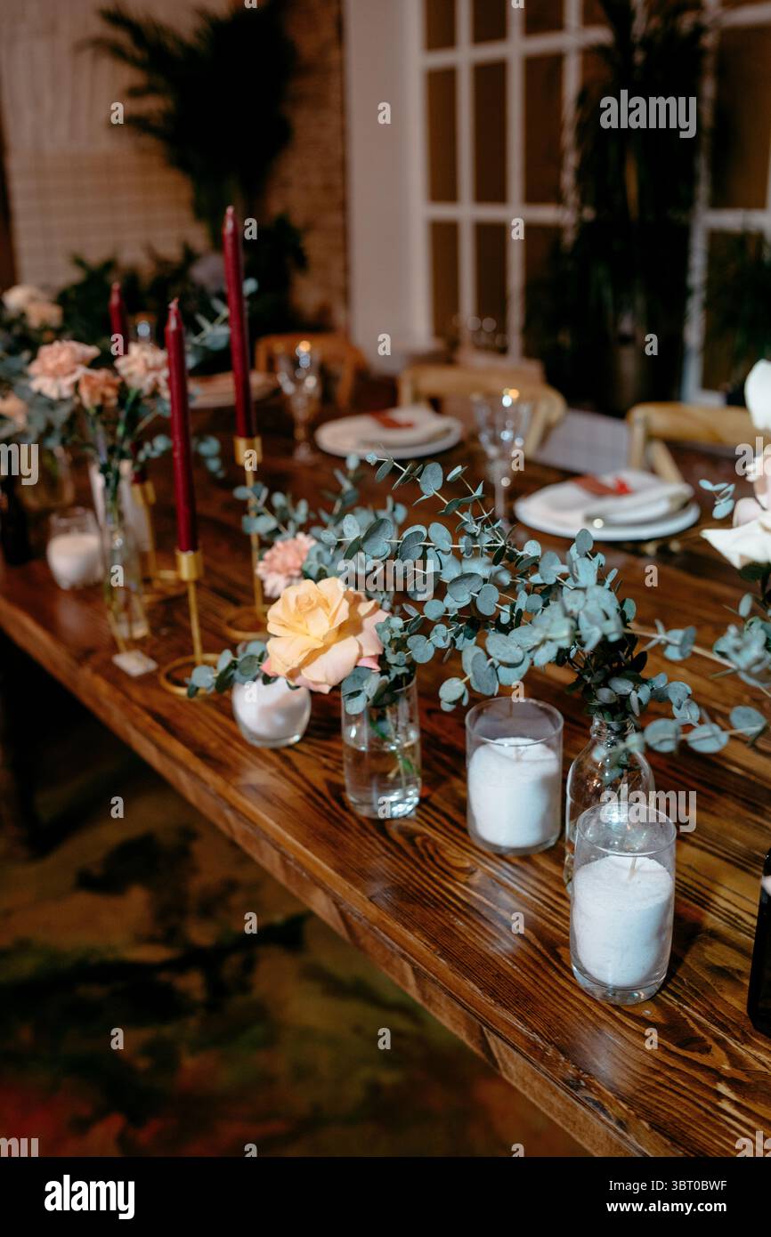 Elegante allestimento da pranzo con candele e composizioni floreali in vasi di vetro su un tavolo di legno, creando un'atmosfera accogliente e sofisticata per un'atmosfera speciale Foto Stock
