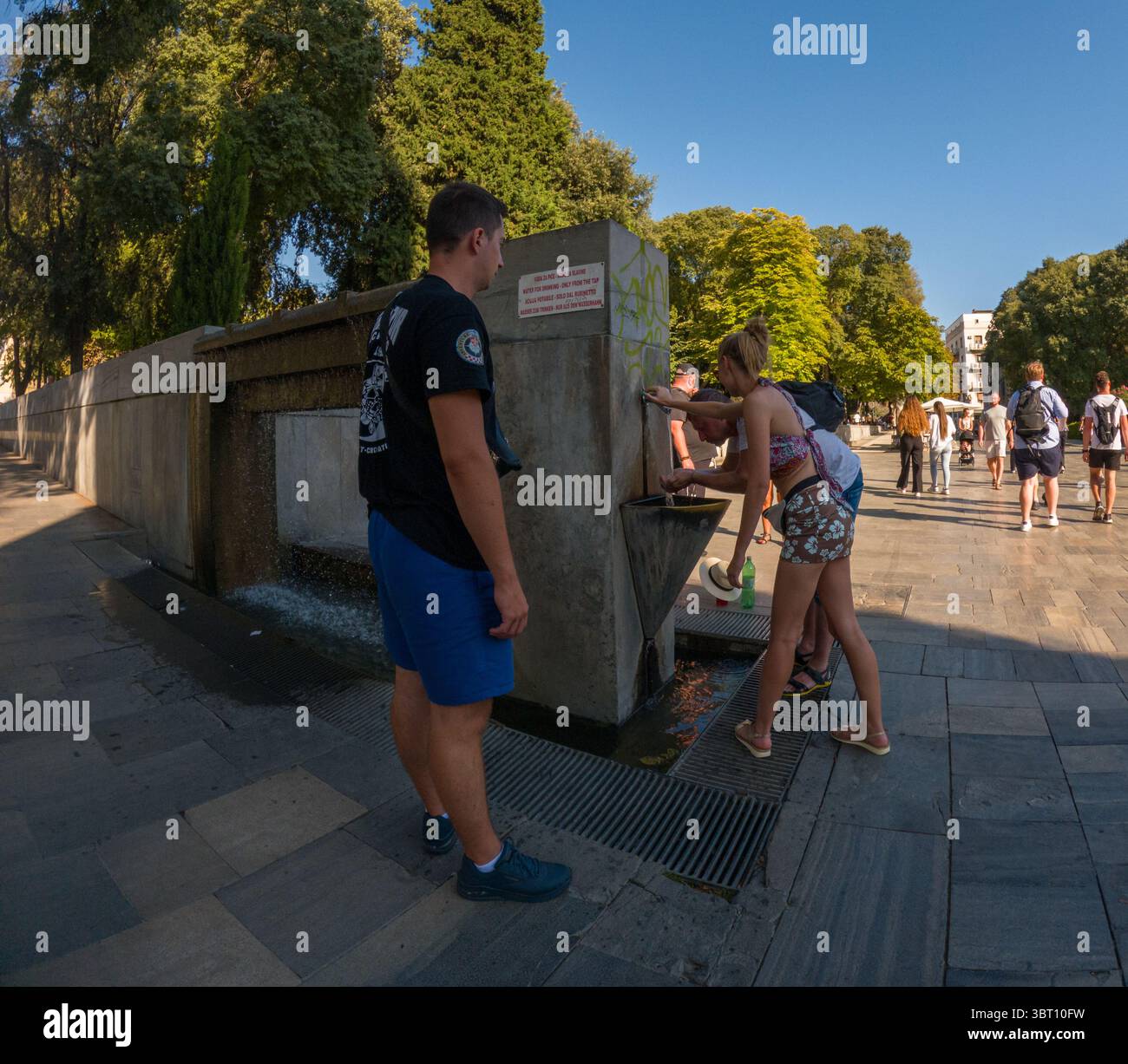 Spalato, Croazia - 8 settembre 2023: I turisti si riuniscono intorno a una fontana pubblica in una giornata di sole, dissetandosi e riempiendo bottiglie d'acqua Foto Stock