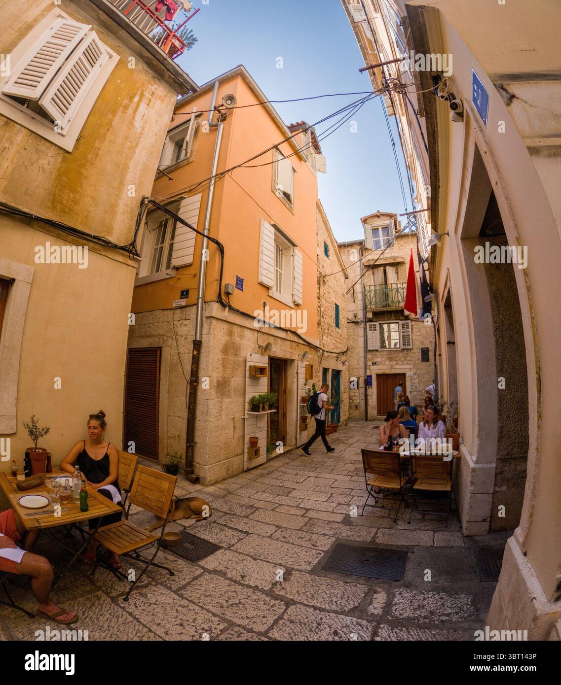 Spalato, Croazia - 8 settembre 2023: La gente si sta godendo un pasto in un caffè all'aperto in una strada stretta. Un uomo passeggia mentre gli altri cenano, creando un mezzo di vita Foto Stock